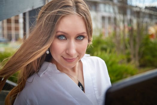 A woman with long hair and captivating blue eyes enjoys a sunny day in a vibrant city atmosphere surrounded by plants.