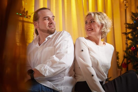 Couple shares a light-hearted moment while sitting beside a decorated Christmas tree in a cozy, festive atmosphere.