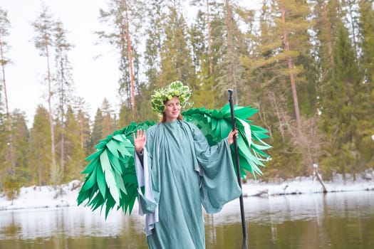 A girl wearing a flowing green dress and flower crown stands by a tranquil lake, embodying the spirit of spring amidst nature.