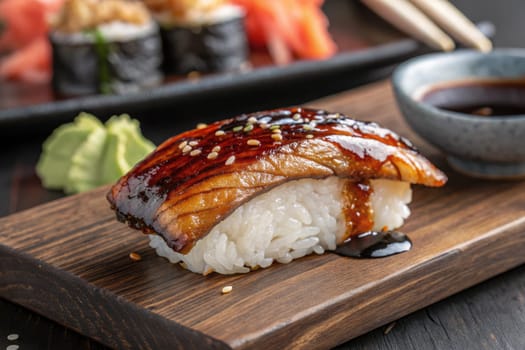 Grilled eel sushi topped with a glossy glaze sits on a wooden plate alongside a portion of wasabi and a small bowl of soy sauce.