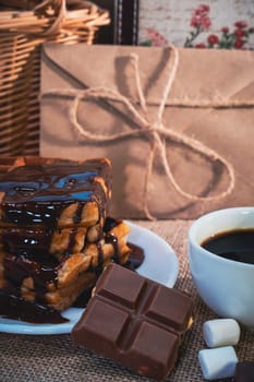 A cup of black coffee with chocolate-covered waffles, a chocolate bar, and marshmallows on a rustic burlap background. Warm and indulgent breakfast or dessert scene
