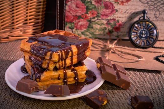 A stack of golden waffles covered in rich dripping chocolate, surrounded by broken chocolate pieces, set on a rustic burlap background with a vintage pocket watch. Warm and indulgent scene