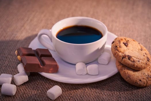 A top-down view of a cup of black coffee with chocolate, a cookie, and marshmallows on a white plate over a textured burlap background. Warm and cozy composition