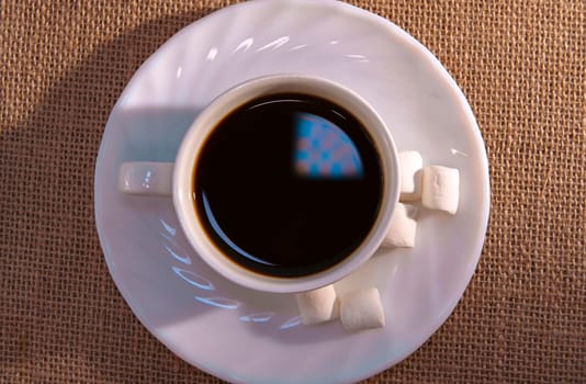 A top-down view of a cup of black coffee with marshmallows on a white plate, placed on a textured burlap background. Warm and minimalistic composition