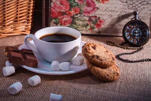 A rustic still life featuring a cup of black coffee, chocolate pieces, cookies, marshmallows, and a pocket watch on a burlap background. Cozy and nostalgic atmosphere
