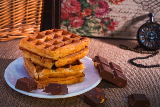 A rustic still life featuring a stack of golden waffles, chocolate pieces with hazelnuts, and a pocket watch on a burlap background. Warm and nostalgic atmosphere