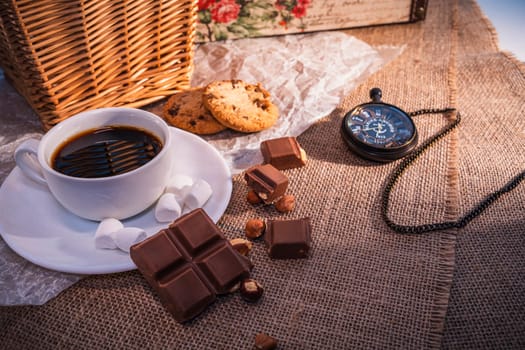 Rustic still life featuring a cup of black coffee, chocolate pieces, cookies, marshmallows, and a pocket watch on a burlap background. Warm and nostalgic atmosphere