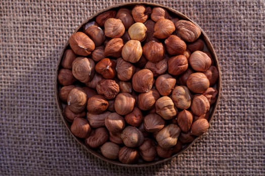 A round bowl filled with whole hazelnuts in their shells, placed on a textured burlap surface. Warm, rustic composition perfect for food and natural themes