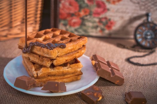 A stack of golden waffles with rich chocolate sauce dripping on top, surrounded by chocolate pieces, set on a white plate with a vintage pocket watch in the background. Warm and indulgent scene