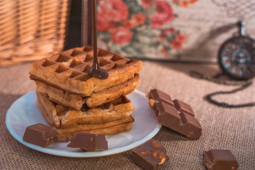 A stack of golden waffles with rich chocolate sauce dripping on top, surrounded by chocolate pieces, set on a white plate with a vintage pocket watch in the background. Warm and indulgent scene