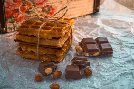 A stack of waffles tied with twine, accompanied by broken chocolate pieces with hazelnuts on crinkled parchment paper. A cozy and nostalgic rustic food composition