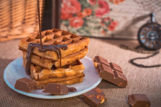 A stack of golden waffles with rich chocolate sauce dripping on top, surrounded by chocolate pieces, set on a white plate with a vintage pocket watch in the background. Warm and indulgent scene