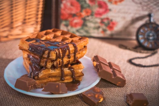 A stack of golden waffles with rich chocolate sauce dripping on top, surrounded by chocolate pieces, set on a white plate with a vintage pocket watch in the background. Warm and indulgent scene
