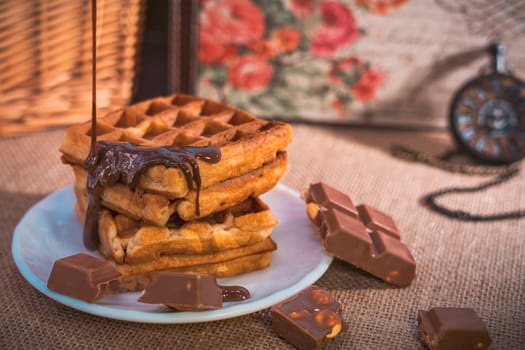 A stack of golden waffles with rich chocolate sauce dripping on top, surrounded by chocolate pieces, set on a white plate with a vintage pocket watch in the background. Warm and indulgent scene