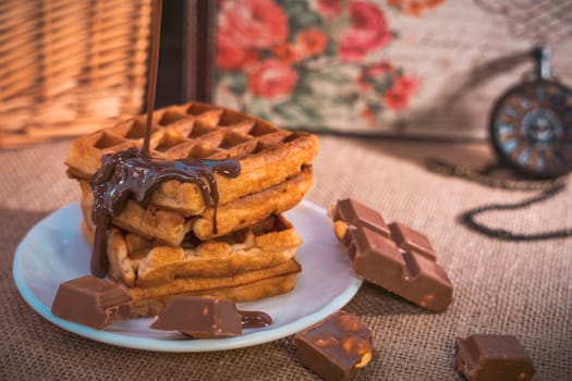 A stack of golden waffles with rich chocolate sauce dripping on top, surrounded by chocolate pieces, set on a white plate with a vintage pocket watch in the background. Warm and indulgent scene