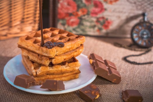 A stack of golden waffles with rich chocolate sauce dripping on top, surrounded by chocolate pieces, set on a white plate with a vintage pocket watch in the background. Warm and indulgent scene