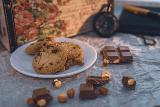 A plate of chocolate chip cookies with broken chocolate pieces and hazelnuts on crinkled parchment paper, accompanied by a vintage pocket watch, notebook, and floral box. Cozy and nostalgic setting