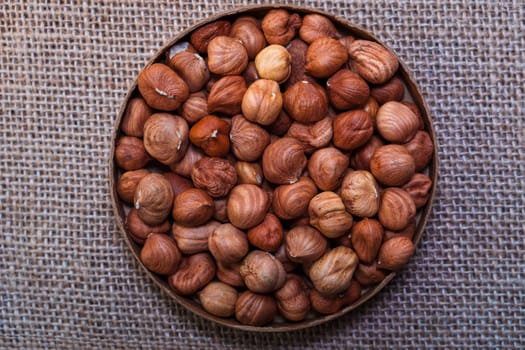 A round bowl filled with whole hazelnuts in their shells, placed on a textured burlap surface. Warm, rustic composition perfect for food and natural themes