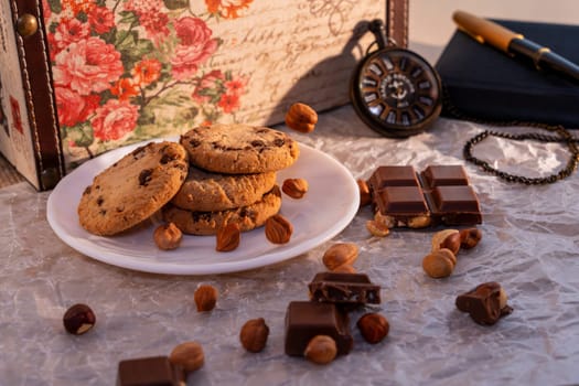 A plate of chocolate chip cookies with broken chocolate pieces and hazelnuts on crinkled parchment paper, accompanied by a vintage pocket watch, notebook, and floral box. Cozy and nostalgic setting