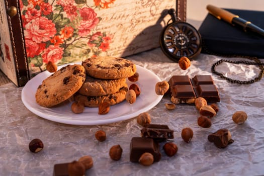 A plate of chocolate chip cookies with broken chocolate pieces and hazelnuts on crinkled parchment paper, accompanied by a vintage pocket watch, notebook, and floral box. Cozy and nostalgic setting