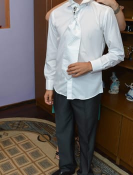 A man dressed in formal attire, wearing a crisp white shirt, a patterned tie, and dark trousers, getting ready for a memorable event with a helping hand in the background.