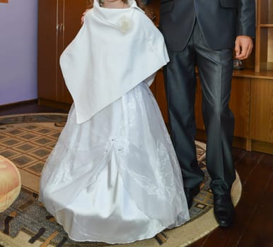A beautifully dressed young girl in a white gown with a stylish cape, accompanied by a formally dressed adult, capturing a special occasion in a warmly decorated indoor setting.