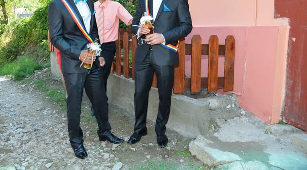Two men in formal suits hold drinks, displaying vibrant sashes and stylish shoes, embodying the spirit of celebration.