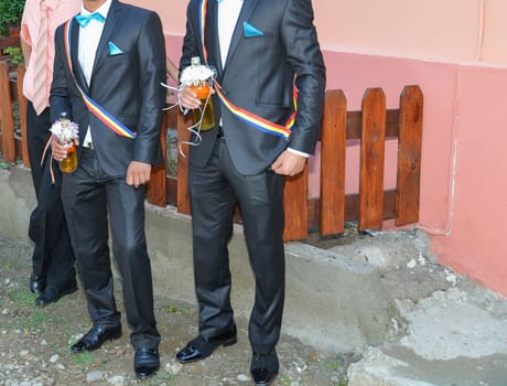 Two men in formal suits hold drinks, displaying vibrant sashes and stylish shoes, embodying the spirit of celebration.