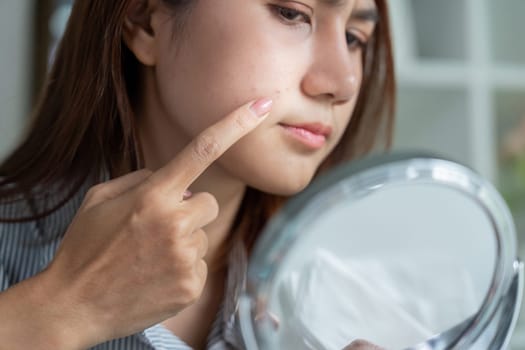 A close-up of a woman examining her skin in the mirror, pointing at a blemish, reflecting the challenges of her beauty regime.