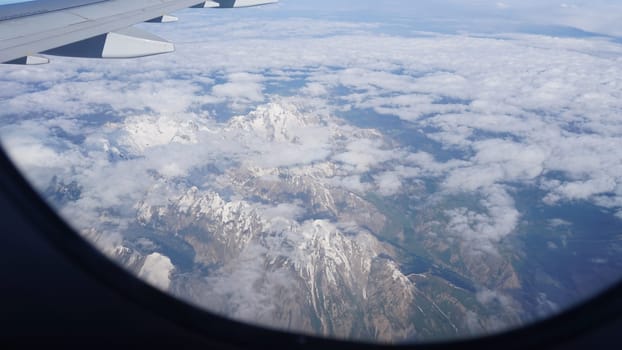 Breathtaking aerial view from an airplane window revealing a majestic mountain range adorned with snow-capped peaks, surrounded by fluffy white clouds drifting across a vibrant sky