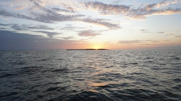 Casting a warm glow, the setting sun illuminates a tranquil ocean, while a tropical island stands silhouetted against a vibrant, colorful sky in the maldives