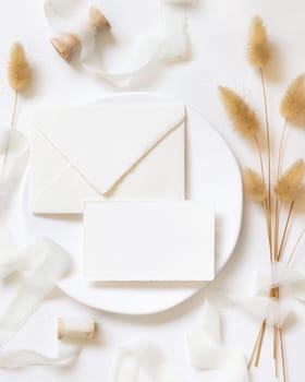 Blank card and envelope on white plate near curled silk ribbons and dried hare's tail grass topview, mockup. Pastel romantic flatlay. Bohemian or close to nature cards for party design or wedding stationery