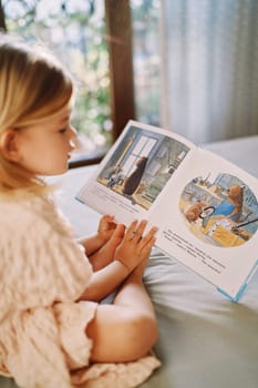 Little girl reads a picture book about a beaver workshop while sitting on her bed. Side view. High quality photo