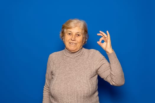 Close-up photo of smiling Caucasian senior retired woman standing casually, showing Ok gesture symbol, looking at camera isolated over blue color background, studio portrait.