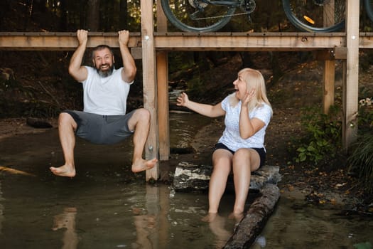 Two friends are having fun by the water on a sunny day. One is playfully hanging from a wooden platform while the other sits on a log, both smiling and enjoying the moment in nature.
