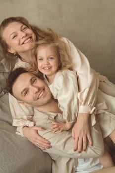 A joyful family of three enjoys a tender moment in a soft-toned indoor space. They are embracing each other with smiles, radiating affection and happiness together.