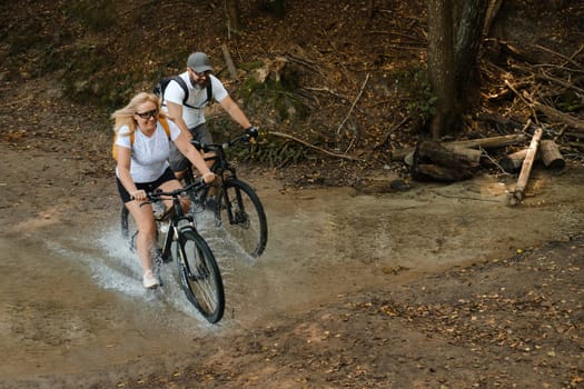 Cyclists navigate through a muddy trail, splashing water as they ride. The wooded area surrounds them, creating a serene environment on a sunny summer afternoon.