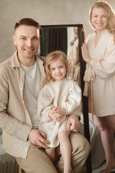 A smiling family poses together in a bright room, with the father holding their daughter while the mother stands beside them. The warm atmosphere highlights their joy and connection.