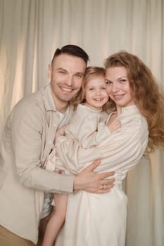 A happy family of three stands closely together, smiling warmly. The parents embrace their child, who is cheerful and playful. Soft lighting creates a warm atmosphere in the background.