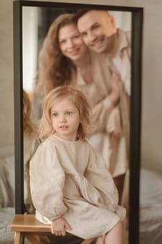 A young girl with long hair sits on a stool in front of a mirror, wearing a light-colored outfit. Her smiling parents are visible in the reflection, creating a warm family atmosphere.