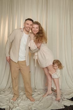 Smiling parents stand close together, embracing their happy toddler who playfully peeks from behind. Soft lighting and neutral colors create a warm, inviting atmosphere in their home.