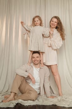 A family of three enjoys a joyful moment indoors, with a child sitting on a parent's shoulders while the other parent stands beside them. The setting features soft drapes and a relaxed atmosphere.