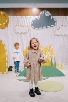 Little laughing girl with a bun in her hand stands on stage among colorful decorations in a kindergarten. High quality photo