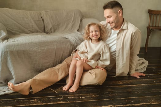 A father sits on the floor with his daughter, both smiling. The cozy living room features soft fabrics and warm tones, creating an inviting atmosphere for their shared time together.
