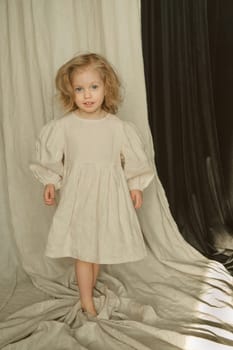 A young girl with curly hair poses in a light dress that contrasts with the softly draped fabrics around her. Natural light enhances the calm atmosphere of the indoor space.