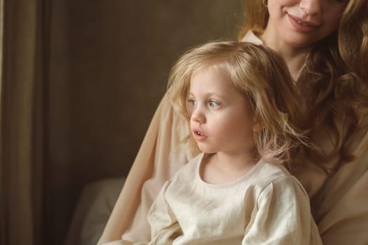 A mother holds her young daughter as they sit together in a warmly lit room. The child gazes thoughtfully, while the mother looks on affectionately, creating a peaceful atmosphere.