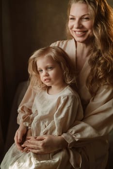 A woman and her young daughter sit together, both smiling. The soft, warm light highlights their expressions, creating a serene and loving atmosphere in the intimate space.