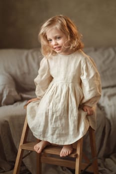 A young girl with wavy hair smiles while sitting barefoot on a wooden stool. Her light-colored dress contrasts with the warm tones of the surrounding cozy space filled with soft materials.