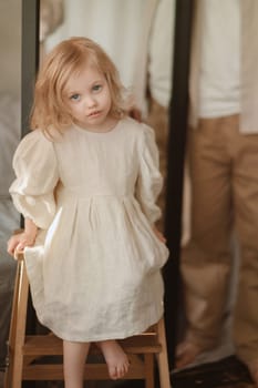 A young girl with wavy blonde hair is seated on a wooden stool, gazing thoughtfully at the camera. The indoor setting is warm and quiet, suggesting an intimate moment in the early afternoon.