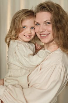 A mother holds her young daughter in a warm embrace, both smiling brightly. The soft lighting creates an intimate atmosphere, showcasing their close connection in a home setting.